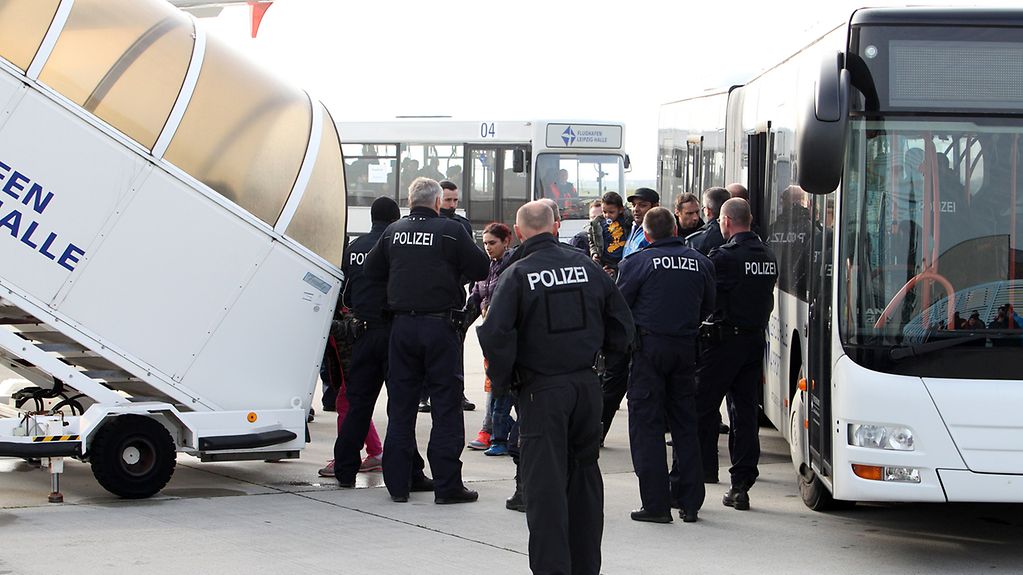Polizisten in weißter Uniform auf einem Flughafen. Im Hintergrund steigen Menschen aus einem Bus aus und betreten über eine Treppe ein Flugzeug.