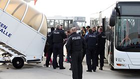 Polizisten in weißter Uniform auf einem Flughafen. Im Hintergrund steigen Menschen aus einem Bus aus und betreten über eine Treppe ein Flugzeug.