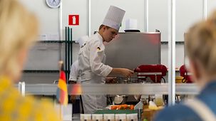 Ein junger Koch mit Kochmütze und weißer Kochuniform hantiert in einer chromfarbenen Küche, im Vordergrund stehen Leute und schauen zu.
