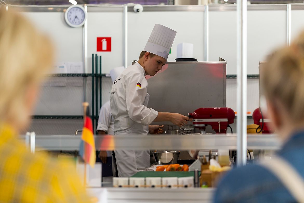 Ein junger Koch mit Kochmütze und weißer Kochuniform hantiert in einer chromfarbenen Küche, im Vordergrund stehen Leute und schauen zu.