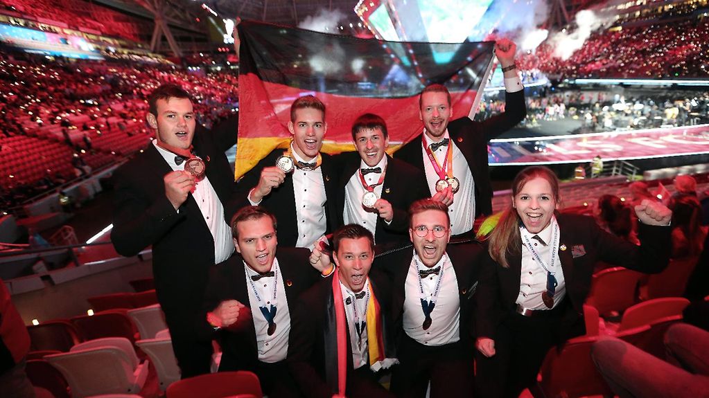 Acht junge Menschen in einem Stadion reißen in Siegerpose die Arme in die Höhe und rufen in die Kamera, während sie eine Deutschlandflagge in den Händen halten.