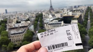 Eine Hand hält eine Eintrittskarte für den Arc de Triomphe - im Hintergrund der Eiffelturm