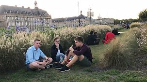 Drei junge Männer machen ein Picknick auf einer Wiese - im Hintergrund historische Gebäude