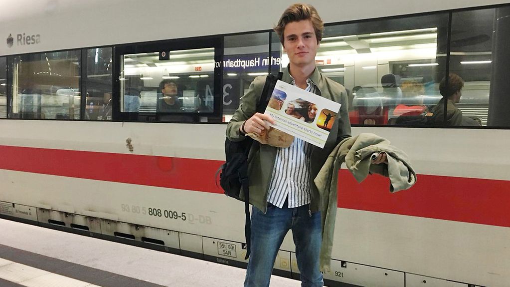 Ein junger Mann steht vor einem ICE am Gleis und hält ein DinA4-großes Ticket in die Kamera.