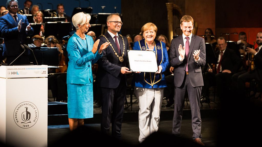 Rector Stephan Stubner presents the Chancellor with a certificate in Leipzig's opera house, while state premier Michael Kretschmar and Christine Lagarde applaud.