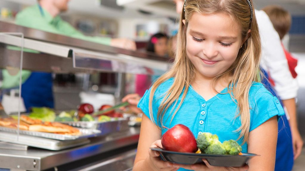 Ein kleines blondes Mädchen steht neben der Essensausgabe einer Schulkantine und hält einen Teller mit Brokkoli und einen Apfel in den Händen-