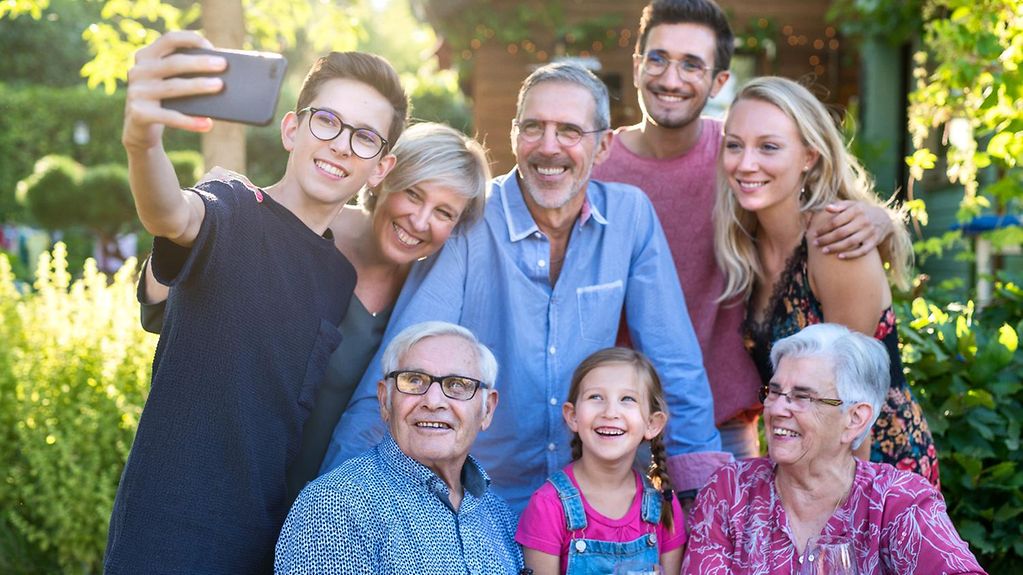 Eine Familie mit Oma, Opa, Eltern und Kindern posiert für ein Selfie und lacht in die Handykamera.