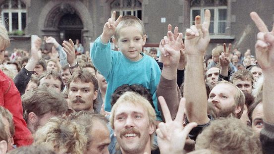 Ein kleiner Junge sitzt auf den Schultern seines Vaters, umringt von einer Menschentraube. Alle recken die Hände zum Siegeszeichen in die Höhe.