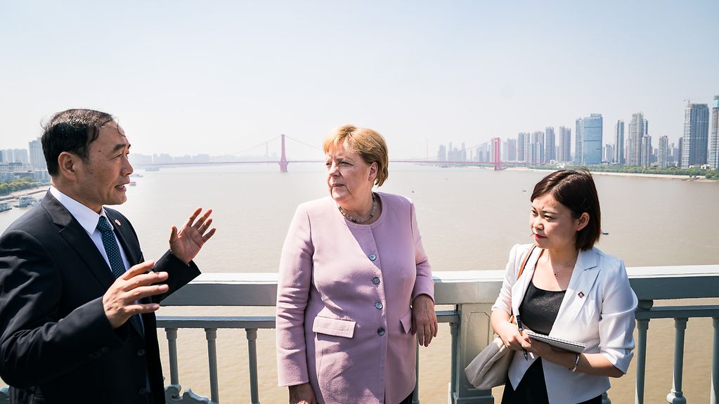 Merkel im fliederfarbenen Blazer auf einer Brücke über einem braunen breiten Fluss, neben ihr ein gestikulierender chinesischer Mann und eine kleine chinesische Frau mit Unterlagen auf dem Arm..