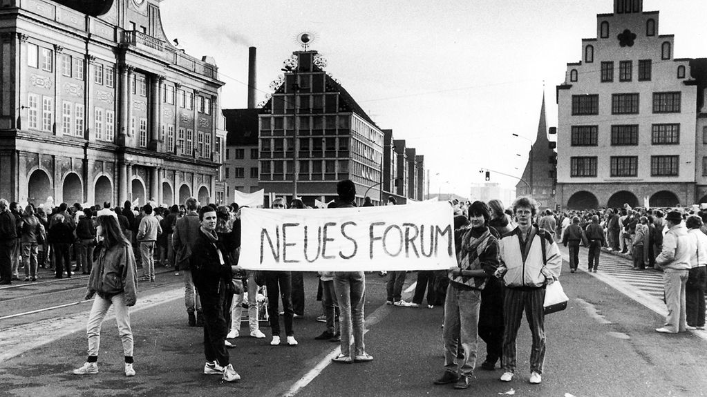 Schwarzweißaufnahme einer Demonstration in Rostock. Die jungen Menschen halten ein Transparent mit der Aufschrift "Neues Forum" in die Kamera.