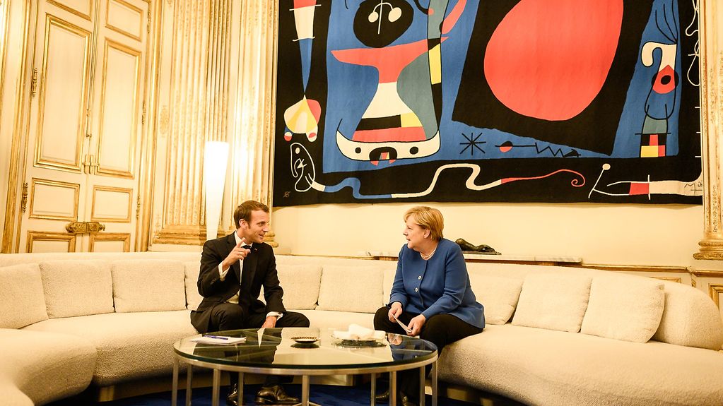 Präsident Emmanuel Macron und Bundeskanzlerin Angela Merkel im Gespräch auf einer hellen, runden Wohnlandschaft. Im Hintergrund ein farbenfrohes Gemälde.