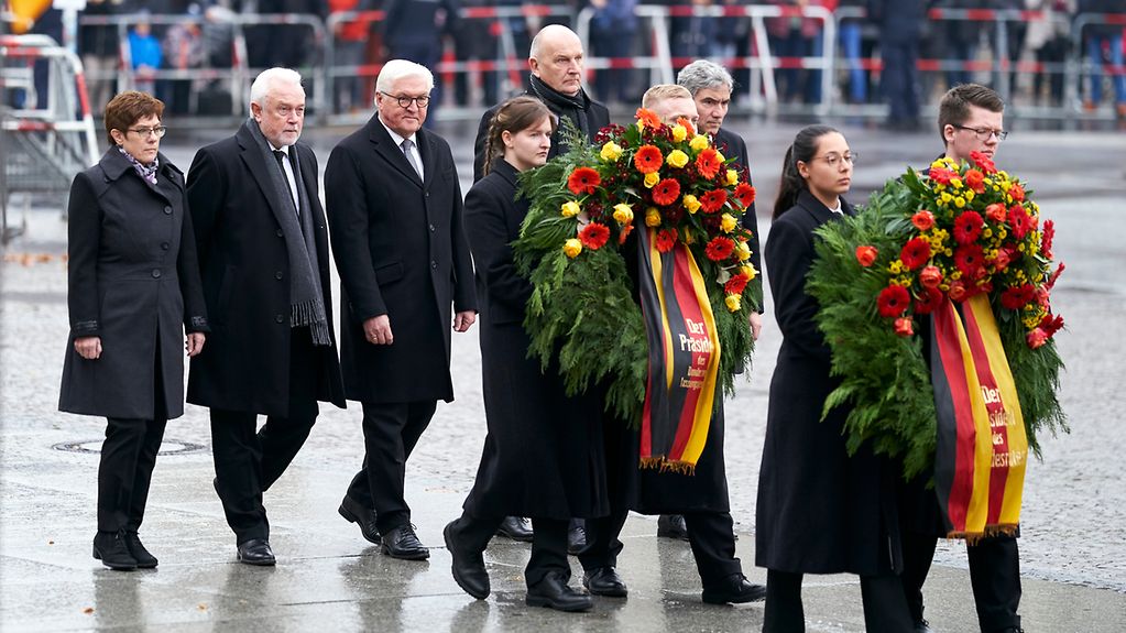 Verteidigungsministerin Annegret-Kramp-Karrenbauer und Bundespräsident Frank-Walter Steinmeier mit eine kleinen Gruppe von Menschen und zwei Blumenkränzen.