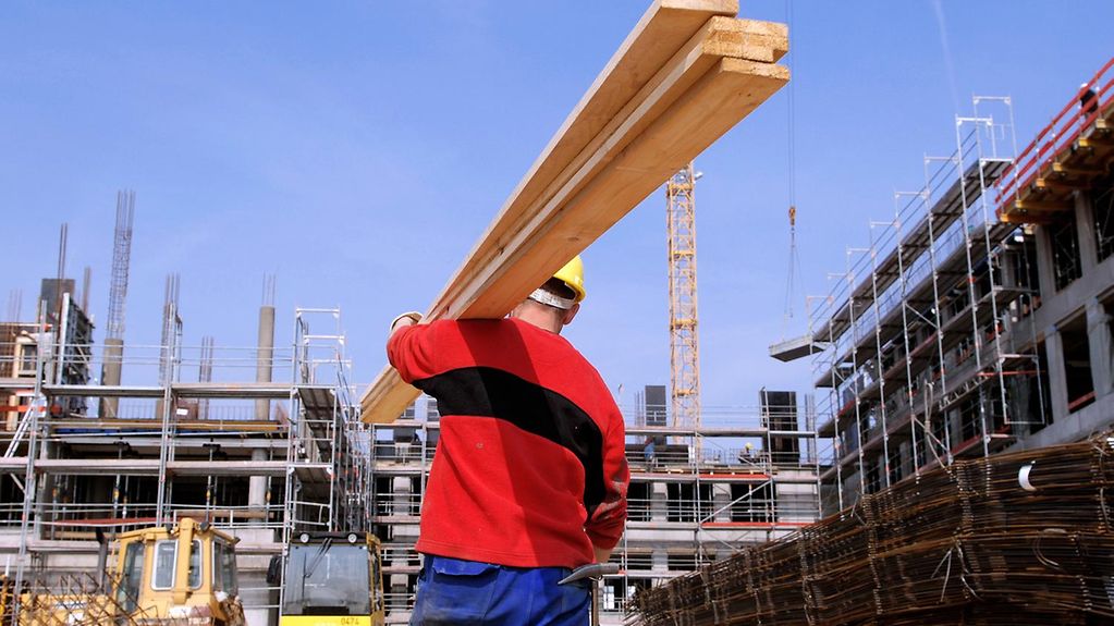 Ein Bauarbeiter mit blauer Schutzhose und Helm trägt eine Holzlatte auf der Schulter und läuft über ein Baustellengelände.