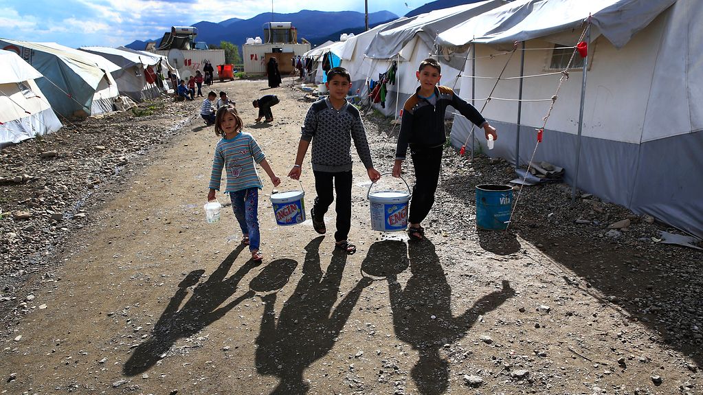 Three people carry buckets of water in a refugee camp.