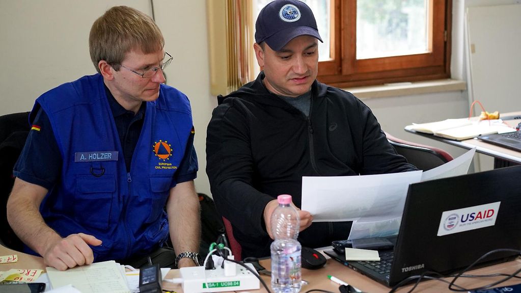 THW-Helfer Achill Holzer bei seinem Einsatz in Albanien, sitzt gemeinsam mit einem weiteren internationalen Helfer an einem Tisch und schaut auf einen Laptop.