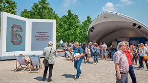 Visitors to the constitutional celebrations beside a large sign saying "Grundgesetz" or German Basic Law. Some visitors are enjoying the summery weather in deck chairs.