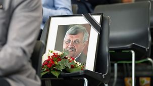 A photo of the deceased with a black ribbon and flowers sitting on an empty chair