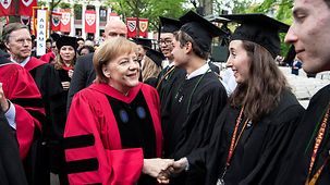 Chancellor Angela Merkel, in a red gown, congratulates relaxed and happy students in academic dress. In the background a number of standards fly.