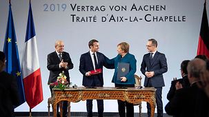 President Emmanuel Macron and Chancellor Angela Merkel at a table, behind them on the wall "2019 Treaty of Aachen". They are flanked by the German and French foreign affairs ministers Heiko Maas and Jean-Yves Le Drian. 