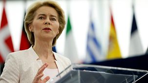 Ursula von der Leyen at the lectern. In the background several indistinct flags of member states can be seen. 