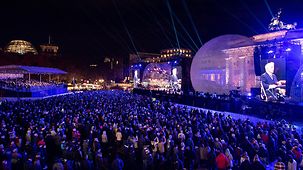 A huge crowd in front of the Brandenburg Gate watch the evening performance. The stage and the musicians on it are bathed in blue light.