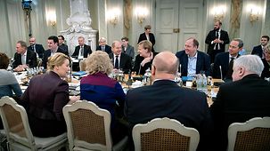 Relaxed Cabinet ministers in lively conversation around a table. Some ministers are seen from behind. In the background other individuals listen.