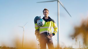 A man in a yellow high vis jacket stands in a field full of wind turbines. He is carrying a helmet and ropes.