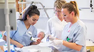 Two trainee nurses receive instruction from a fully qualified nurse in a patient's room. In the background a patient lies in bed.