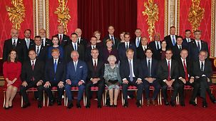 Family photo in a room decorated in red. Her Majesty the Queen is surrounded by the leaders. The front row is seated, while the back rows stand. 