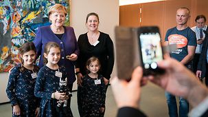 Someone takes a photo of the Chancellor with a phone. She is surrounded by three children and a woman. In the background hangs an abstract picture.