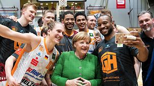 Chancellor Angela Merkel surrounded by basketball players on the court. Selfies are being taken.