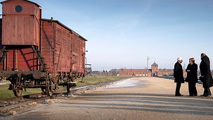The Chancellor talks to two people in front of the entrance to the concentration camp. At the left hand side stands an old railway car on rails.