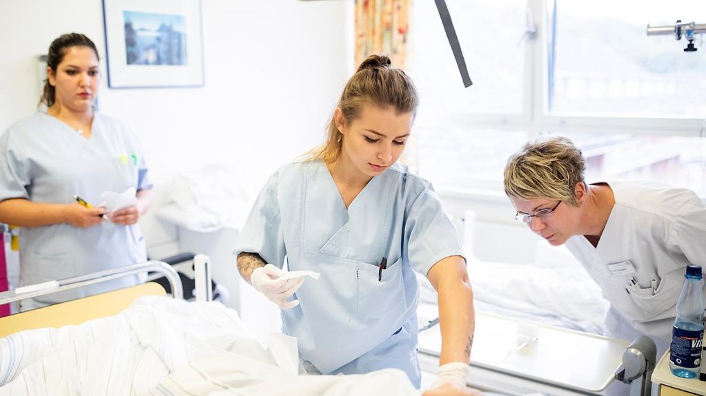 Eine junge Pflegefachkraft beugt sich über ein Krankenhausbett, beobachtet von einer Krankenhausschwester mittleren Alters. Im Hintergrund steht eine weitere junge Azubine.