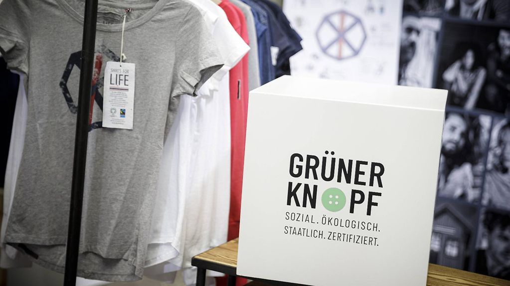 T-Shirts an einem Kleiderständer und ein schild mit dem Label "Grüner Knopf"