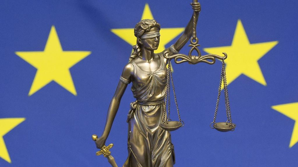 Bronzene Figur der römischen Göttin der Gerechtigkeit, Justitia. Die Figur trägt ein Schwert und eine Waage in den Händen. Im Hintergrund ist die EU-Flagge zu sehen.