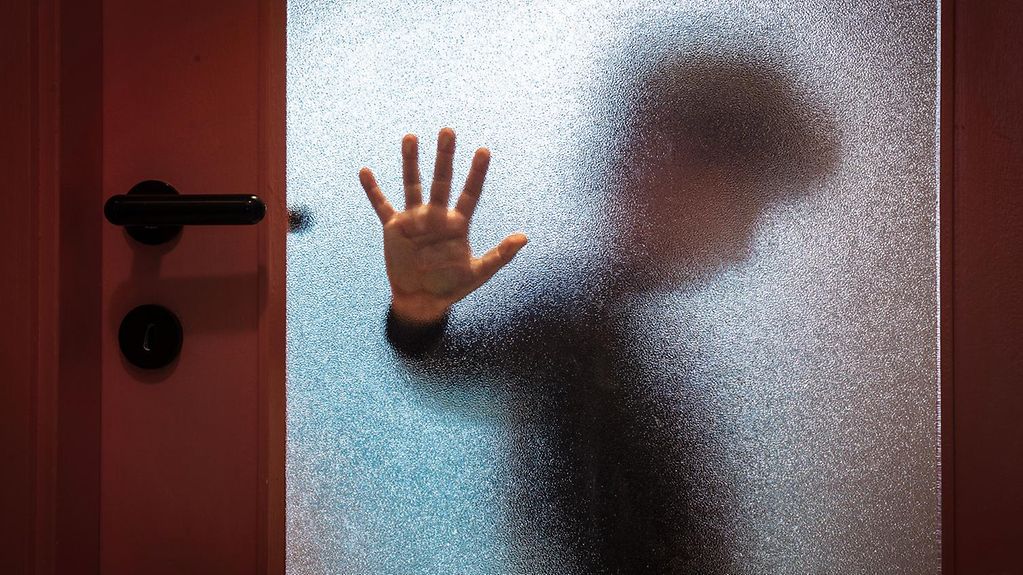Hinter einer Glastür erkennt man schemenhaft ein Kind, das die Hand gegen die Scheibe drückt.