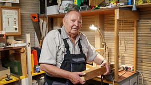 Hiorst Sickert steht an seiner Werkbank und zeigt ein Stück Holz, an dem er gerade arbeitet.
