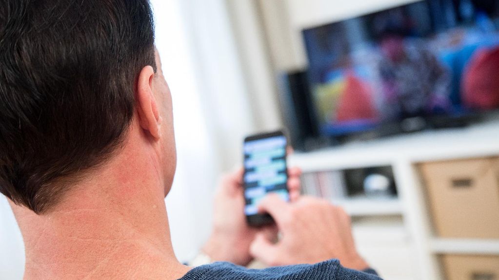 Ein Mann sitzt mit einem Smartphone in der Hand vor einem Fernseher.