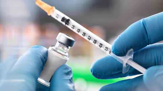 Eine Hand in medizinischen Einweg-Handschuhen hält eine Spritze und einen kleinen Glasbehälter mit Impfstoff.