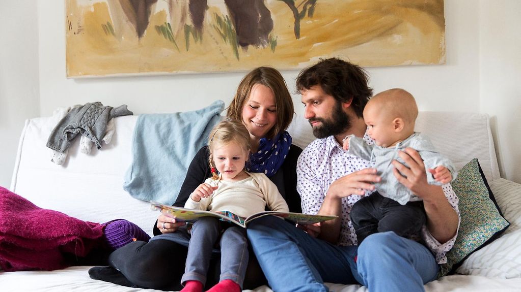 Eltern sitzen mit zwei Kindern auf der Couch und lesen ein Buch.