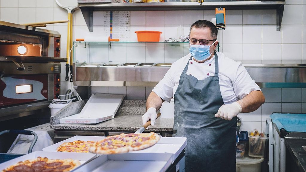 Das Bild zeigt einen Pizzabäcker, der Pizzen aus dem Ofen holt und einen Mundschutz trägt.