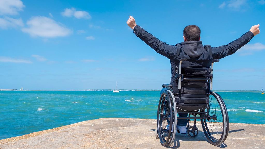 Ein Rollstuhlfahrer reißt mit Blick auf das Meer die Hände hoch.