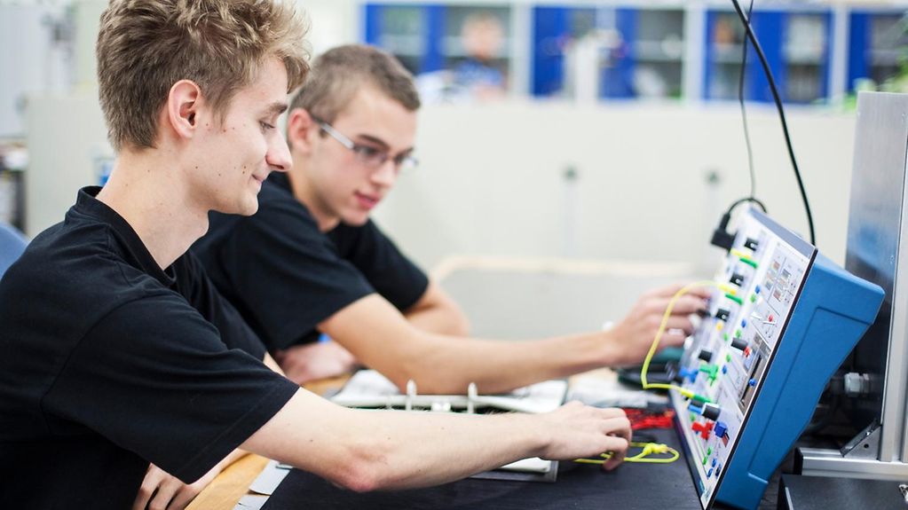 Zwei junge Auszubildende zum Elektroniker für Betriebstechnik sitzen vor einem Steuergerät.