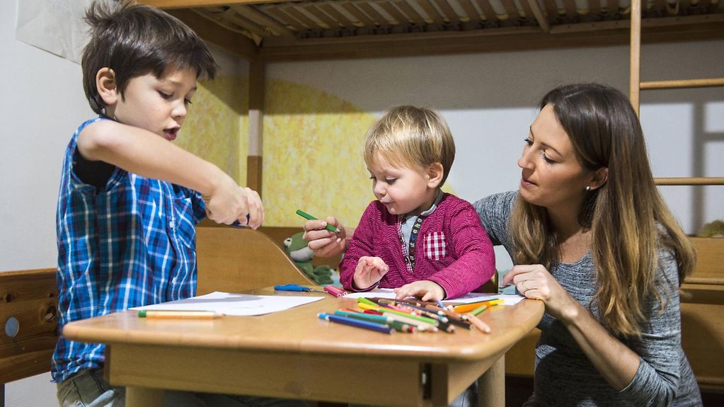 Das Bild zeigt Brüder, die im Kinderzimmer mit Buntstiften spielen, während die Mutter ihnen assistiert.