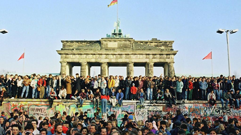 Sehr viele Menschen sitzen und stehen vor der Mauer und auf der Mauer vor dem Brandenburger Tor. 