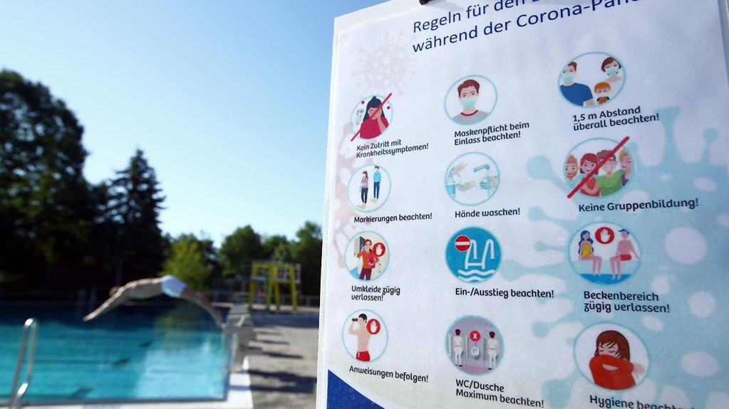 Eine Tafel mit Corona-Verhaltensregeln für das Freibad, im Hintergrund springt ein Mann vom Beckenrand ins Wasser.