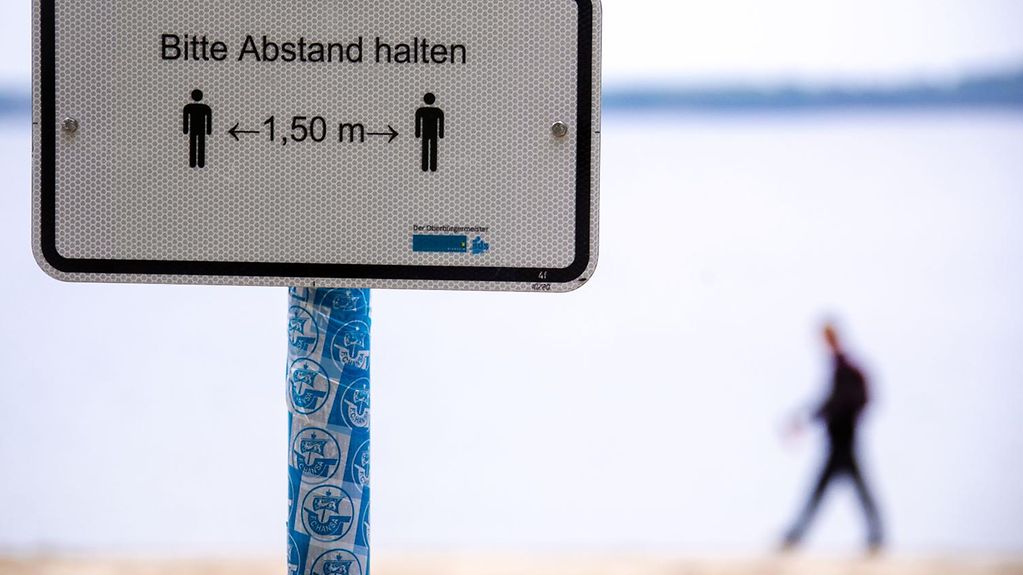 Ein Spaziergänger wandert am Ufer eines Sees entlang. Im Vordergrund steht ein Hinweisschild über den Mindestabstand von 1,5 Metern.