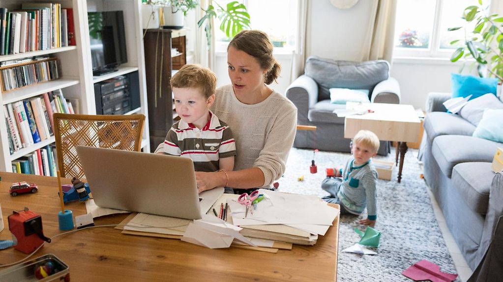 Eine Mutter mit zwei Kindern sitzt am Laptop in ihrem Wohnzimmer.