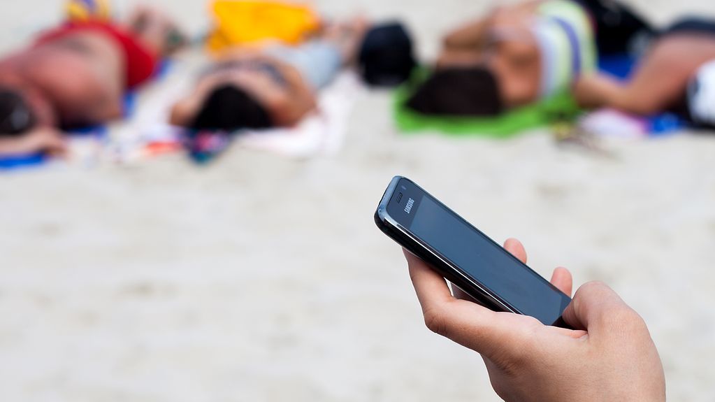 Eine Frau schreibt mit einem Mobiltelefon eine SMS am Strand, im Hintergrund sonnen sich Urlauber auf Handtüchern.