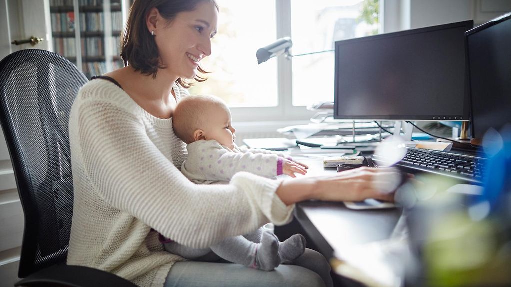 Lächelnde Mutter sitzt am Schreibtisch vorm Computer mit Baby auf dem Schoß.